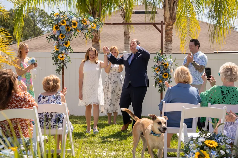 Loved ones gather in white chairs around the ceremony space as the couple stands under the arch with a golden dog at their feet.