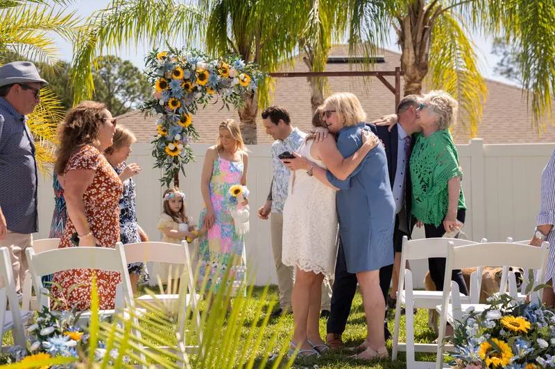 Small groups of guests embrace and celebrate near the arch as the ceremony concludes in the palm-lined yard.