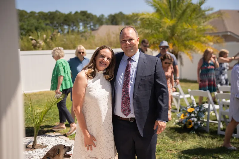 Danielle and Von stand together in a cream and navy suit at their St. Augustine Airbnb ceremony, with guests and tropical plants visible behind them.