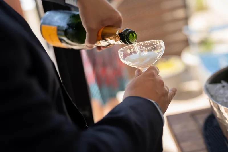 Hands in dark sleeves pour champagne into a coupe glass during a backyard celebration.