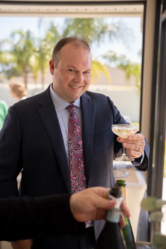Someone in a navy suit and patterned tie raises a champagne glass at an indoor St. Augustine gathering, palm trees visible through windows.