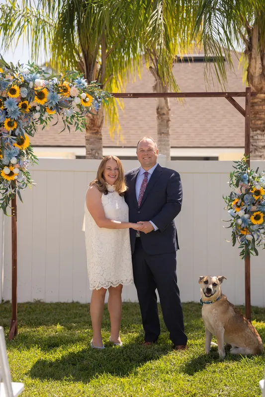 Guests seated around a reflective pond watch as two people stand together under a decorated arch surrounded by palm trees.