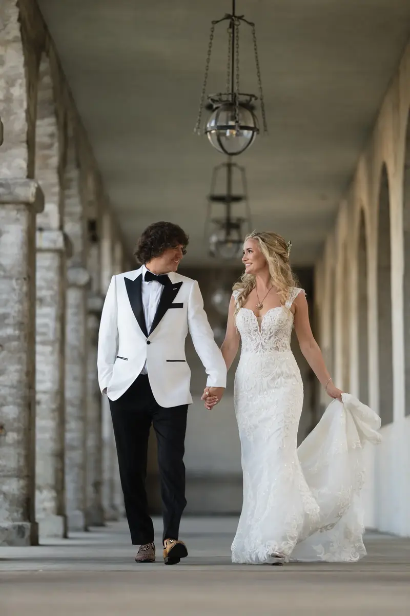 Diana and Liam walk hand in hand down a columned hallway at Lightner Museum, with Diana's white gown trailing behind them.