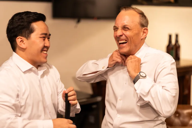 Two men in white dress shirts laugh together while tying neckties indoors.