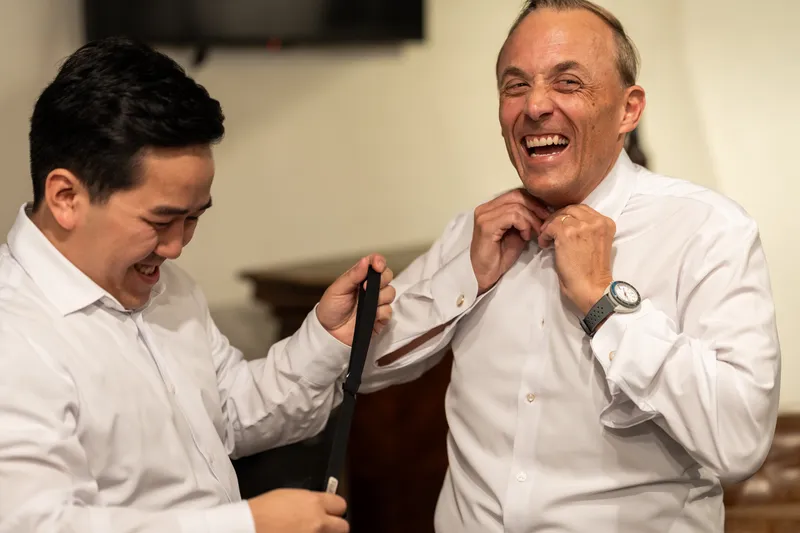 Groom and groomsman laughing while adjusting shirt collar before the ceremony