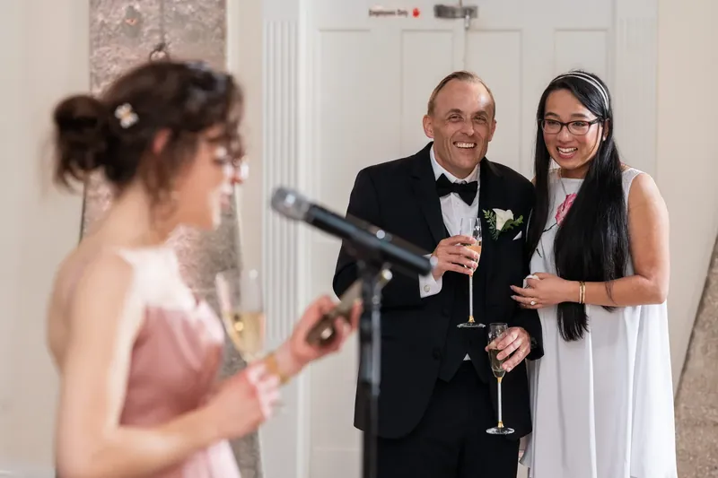A speaker holds a champagne glass as a man and woman in formal wear smile beside her at the reception.