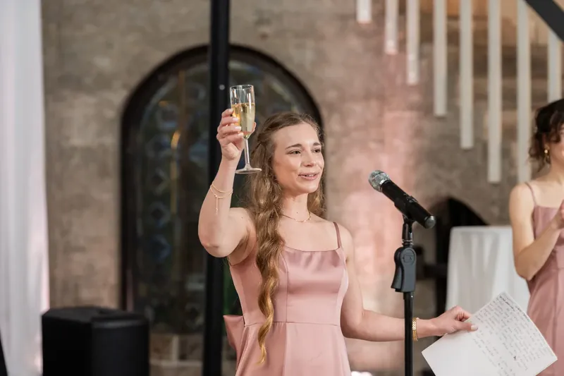 A bridesmaid raises her champagne flute while holding her speech notes, her expression joyful and engaged.