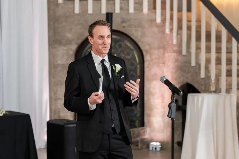 Jason stands at the microphone in his black tuxedo, gesturing expressively while delivering remarks at the reception.