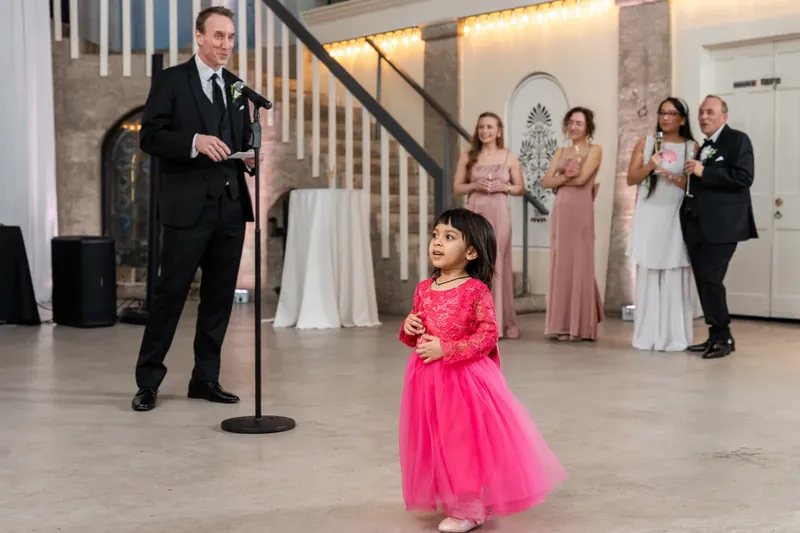 Jason watches a young girl in a bright pink dress dance across the grand hall at Lightner Museum while wedding guests look