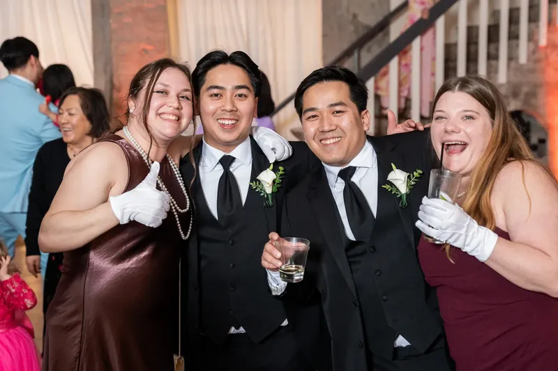 Jason and two groomsmen laugh with guests in formal attire, white boutonnieres pinned to their lapels.