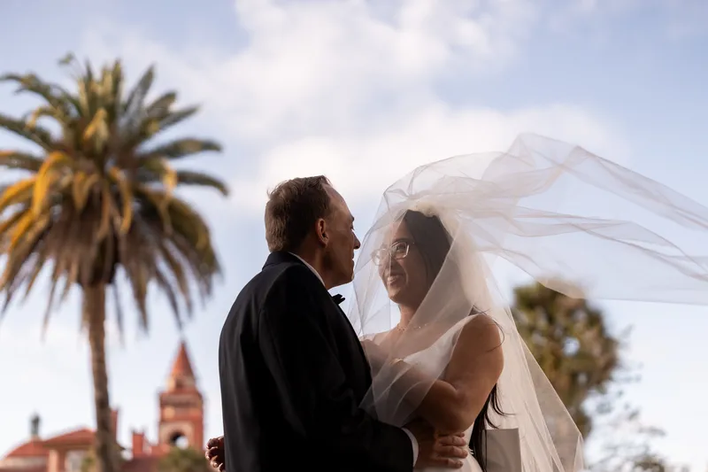 Bride and groom embracing with veil blowing in the wind, palm tree and Flagler College behind them