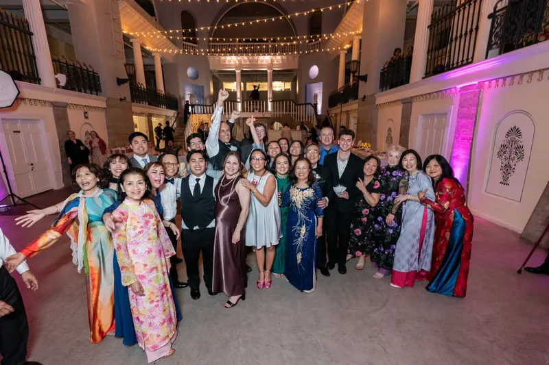 The entire wedding party and family gather for a group portrait under string lights in the Lightner Museum's grand hall.