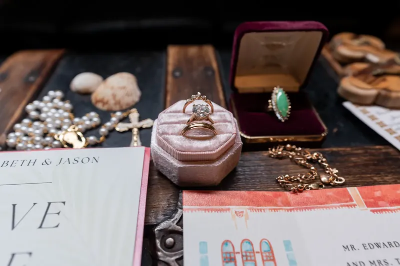 Elizabeth and Jason's rings and jewelry displayed on dark wood with invitation cards nearby.