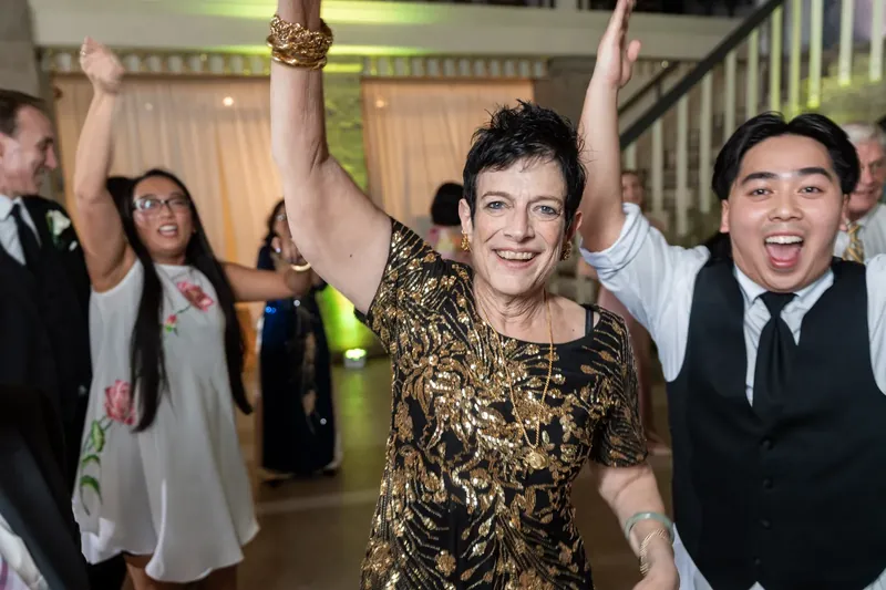Woman in gold sequined dress raises arms celebrating with guests on dance floor at Lightner Museum.