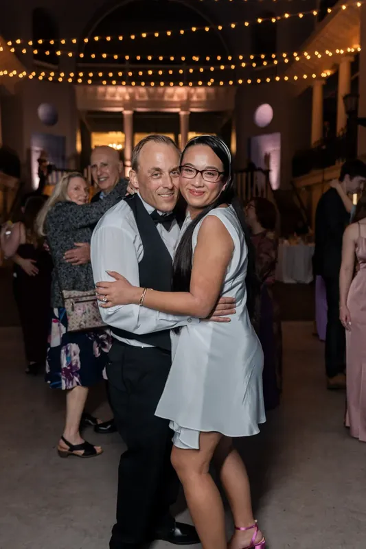 Couple embraces under string lights on dance floor surrounded by guests at Lightner Museum.
