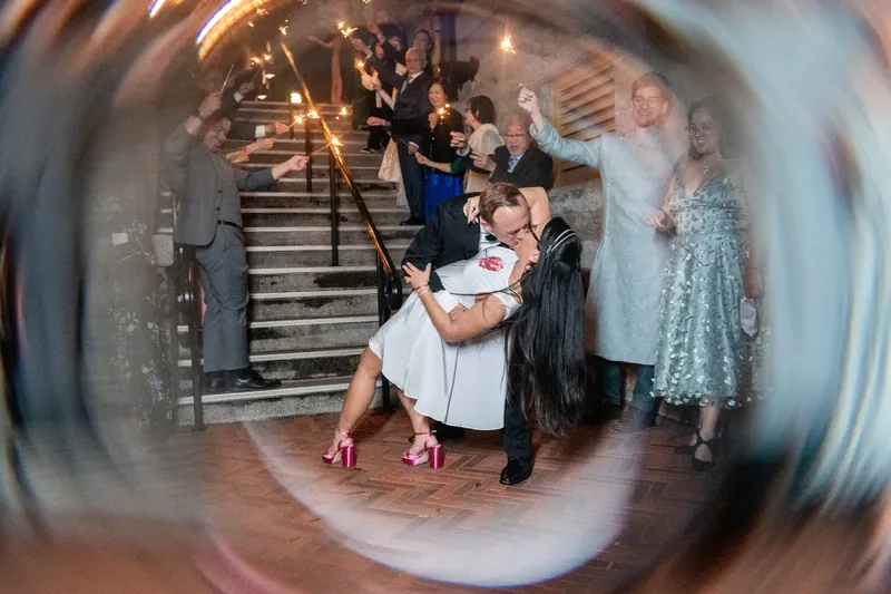 Elizabeth lifts her white dress while Jason supports her on the ornate staircase, captured through a dreamy circular lens.