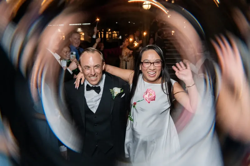 Jason and Elizabeth celebrate at the Lightner Museum, both grinning widely as confetti swirls around them in a dynamic