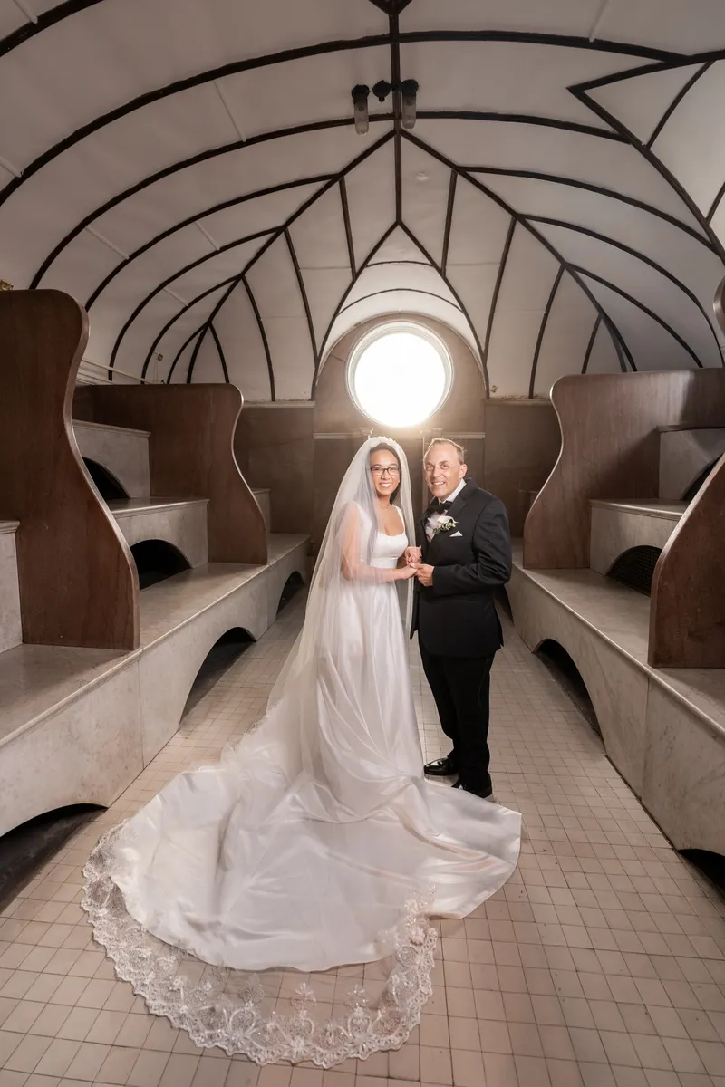 Couple holding hands in the Lightner Museum Russian Baths with cathedral train and veil backlit by round window