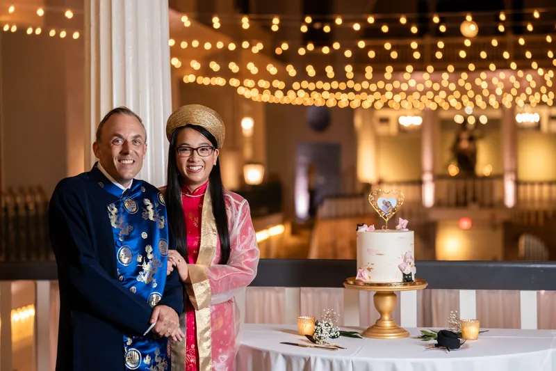 Couple in ao dai posing by wedding cake with string lights in the Lightner Museum ballroom