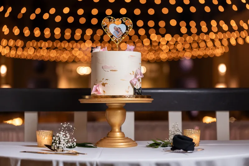 Wedding cake with gold leaf, pink flowers, heart photo topper, and string light bokeh behind
