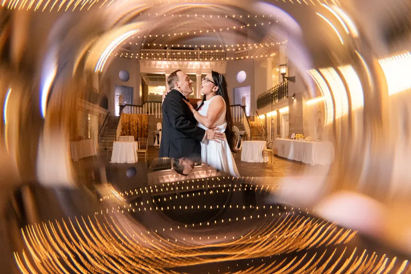 Ring-shot portrait of couple dancing in the empty Lightner Museum ballroom with string lights