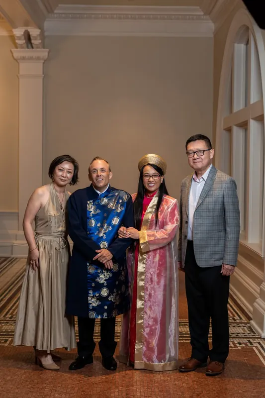 Four adults in traditional South Asian wedding attire stand together at the Lightner Museum, with the men in embroidered silk