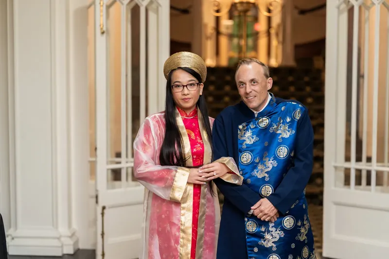 Elizabeth and Jason hold hands while exchanging vows, both wearing richly embroidered blue and pink silk wedding attire