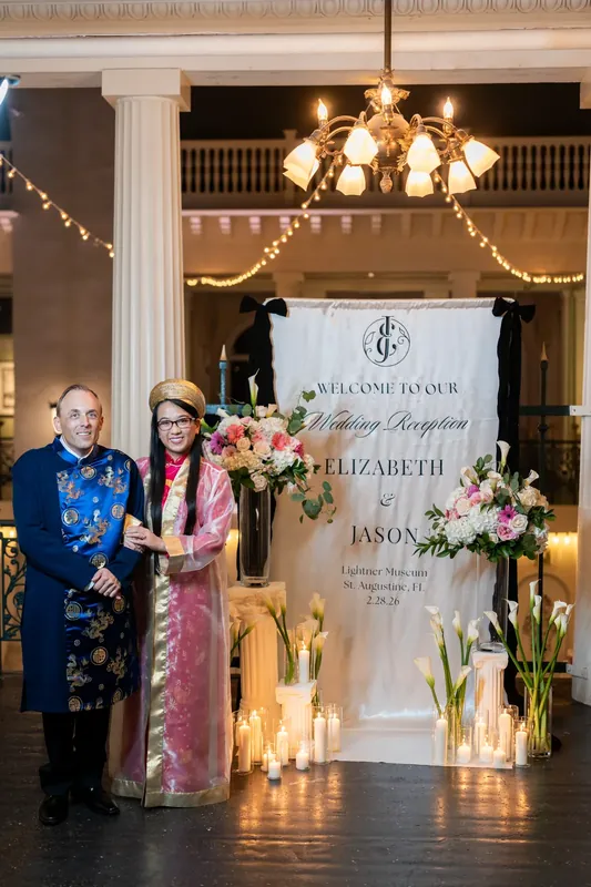 Jason and Elizabeth stand beside an ornate wedding reception sign flanked by floral arrangements and glowing candles beneath
