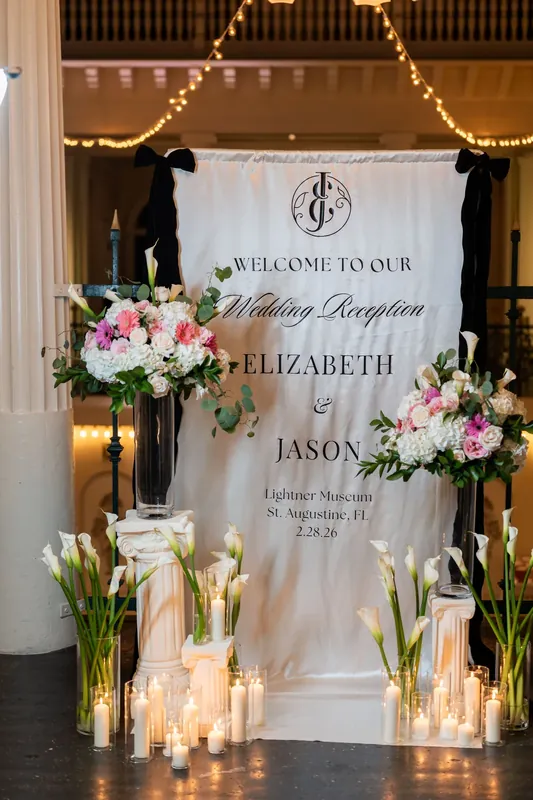 Tall glass candles frame a white wedding reception sign reading "Welcome to Our Wedding Reception" with pink and white floral