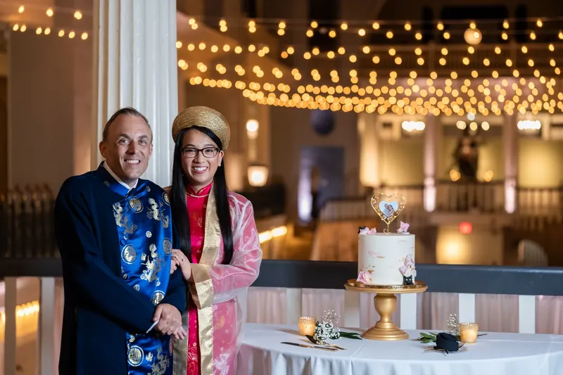 Jason and Elizabeth stand together under glowing string lights, smiling beside their wedding cake.