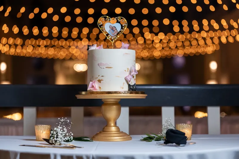 A white tiered cake with purple florals and a heart topper sits on a gold pedestal beneath strung bulbs.