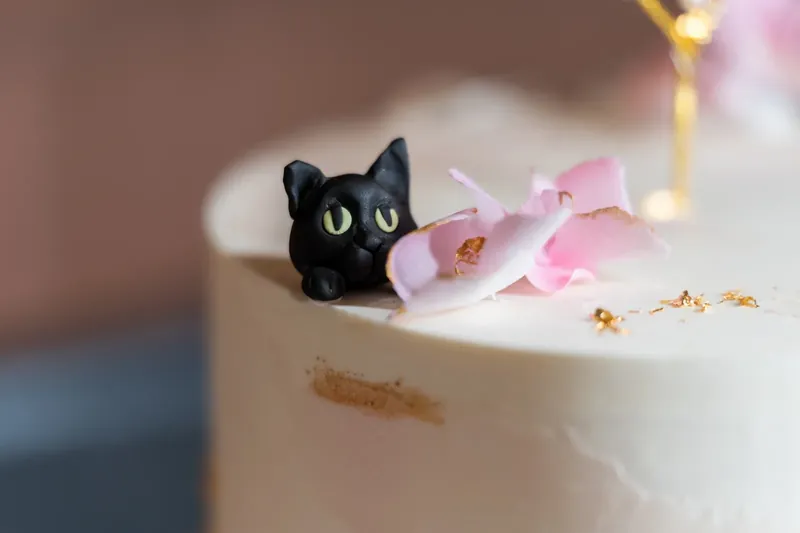 A black cat figurine perches on the cake beside pink orchid petals and gold leaf details.