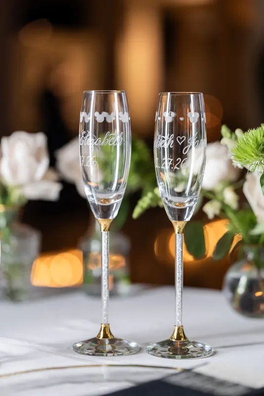 Two champagne flutes engraved with Elizabeth and Jason's names sit side by side among white flowers.