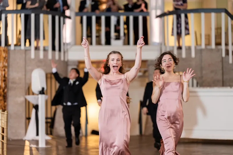 Two bridesmaids in dusty rose dresses leap and cheer during the reception at Lightner Museum, with guests in formal attire