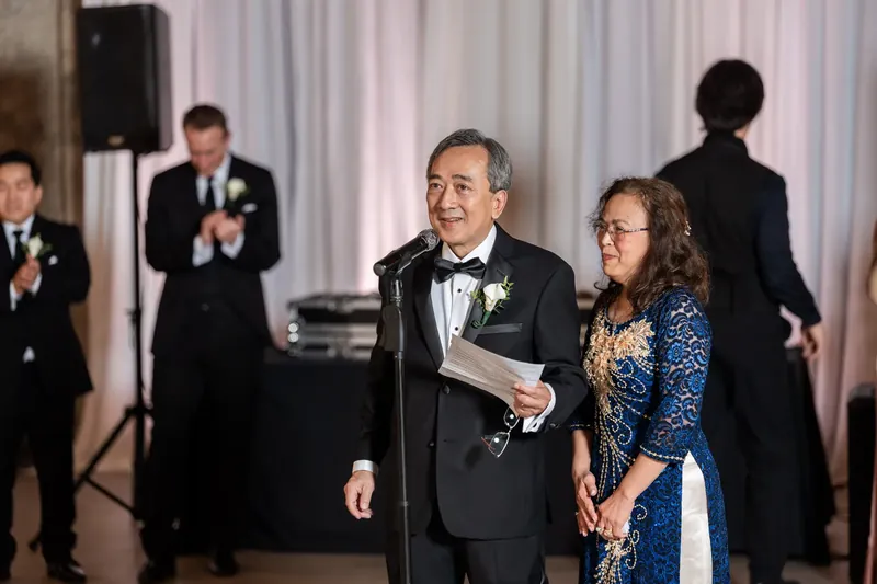 A man in a tuxedo speaks at a microphone while a woman in a blue dress stands beside him at the Lightner Museum.