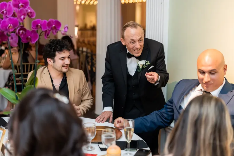 A man in a tuxedo speaks animatedly at a dinner table adorned with bright pink flowers.