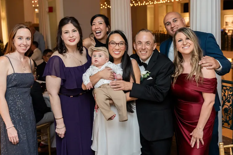 Elizabeth and Jason pose with their families beneath ornate columns at the Lightner Museum, wearing formal attire and holding