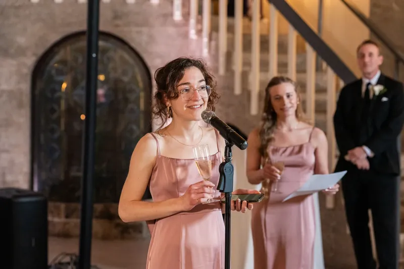 A bridesmaid in blush pink speaks at a microphone while guests listen from the elegant venue.