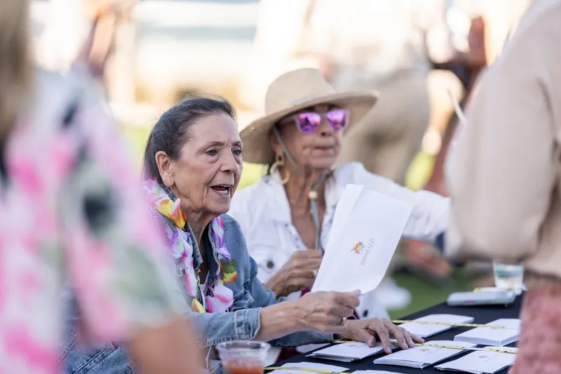 Two women laugh while seated at a table, one wearing a cowboy hat and pink-tinted sunglasses, the other in a colorful scarf.