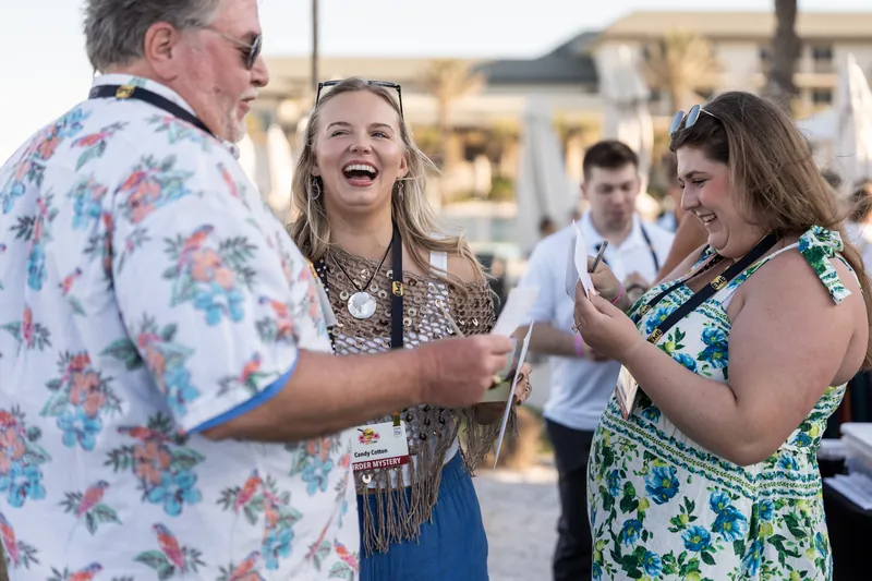A man in a floral shirt chats with two women at an outdoor event, all three smiling and holding drinks.