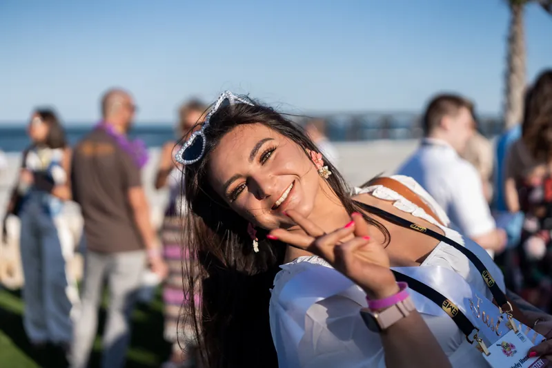 A woman with sunglasses on her head tilts her head playfully toward the camera with ocean and guests visible behind her.