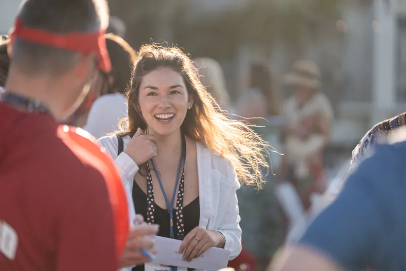 A woman in a white cardigan and beaded necklace grins while conversing with guests at an outdoor gathering.