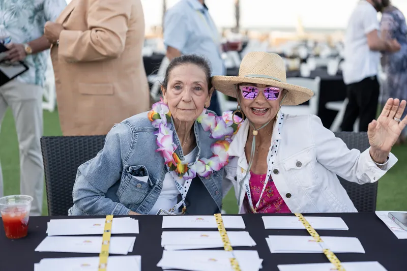 Two people seated at a table wearing lei and sunglasses smile for the camera, surrounded by event attendees.