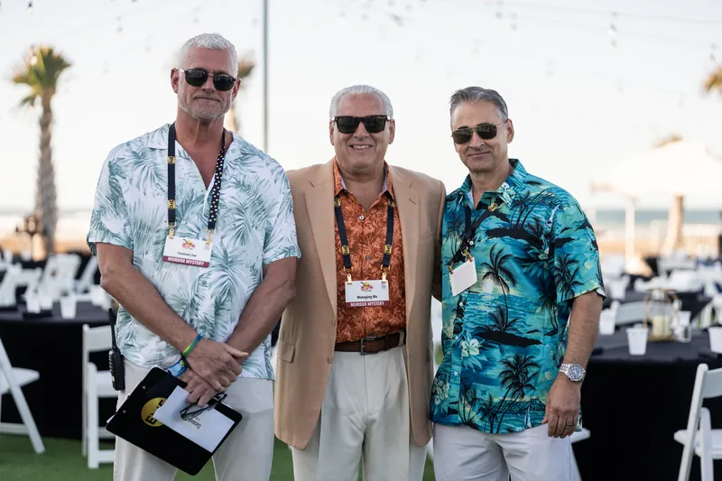 Three men stand together in tropical and floral print shirts against a palm tree and ocean backdrop.
