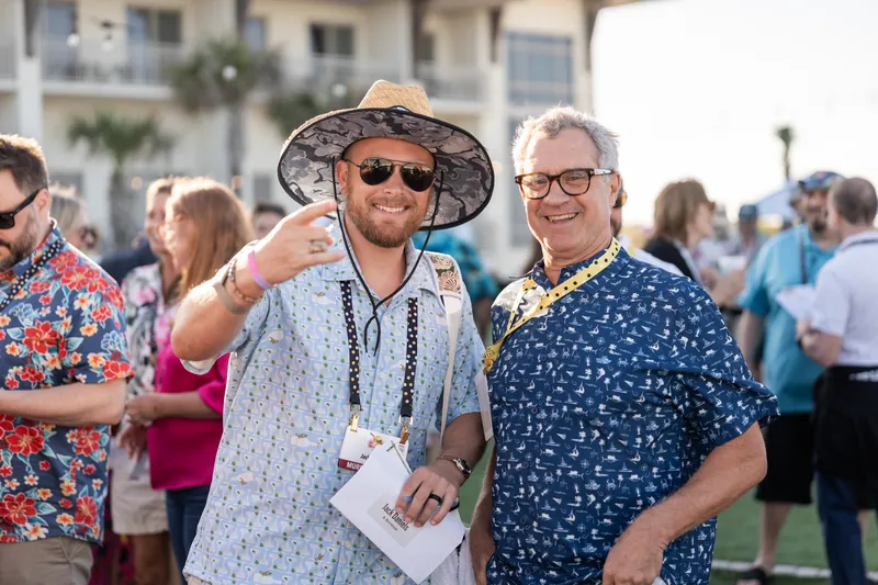 Two men smile at the camera wearing sunglasses and patterned shirts, one in a straw hat, at an outdoor event with guests