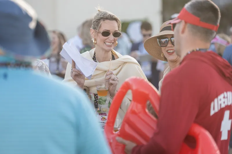 A man in sunglasses and light shirt chats with guests near a pool area, holding a drink and wearing an event credential.