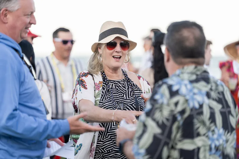 Guests in bright printed fabrics and hats socialize and laugh during the beachfront celebration, with the hotel visible