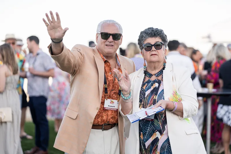 An older man in a tan blazer waves enthusiastically while standing beside a woman in sunglasses and a patterned shirt