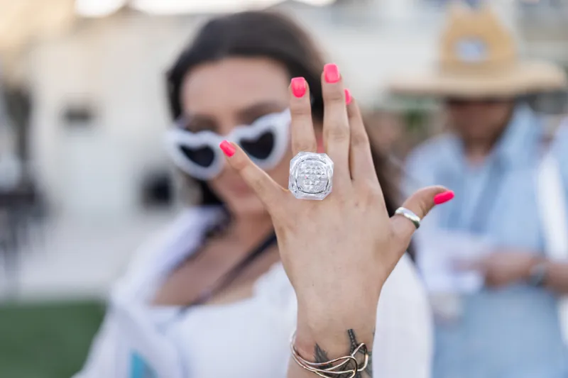 A woman displays her diamond ring and pink manicure while wearing sunglasses at the outdoor event.