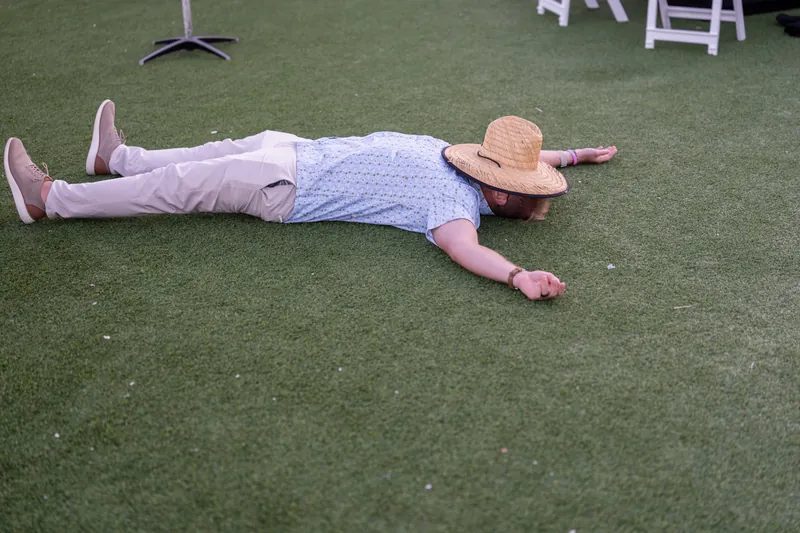 A man in pink and blue sprawls dramatically across the grass with his straw hat beside him.